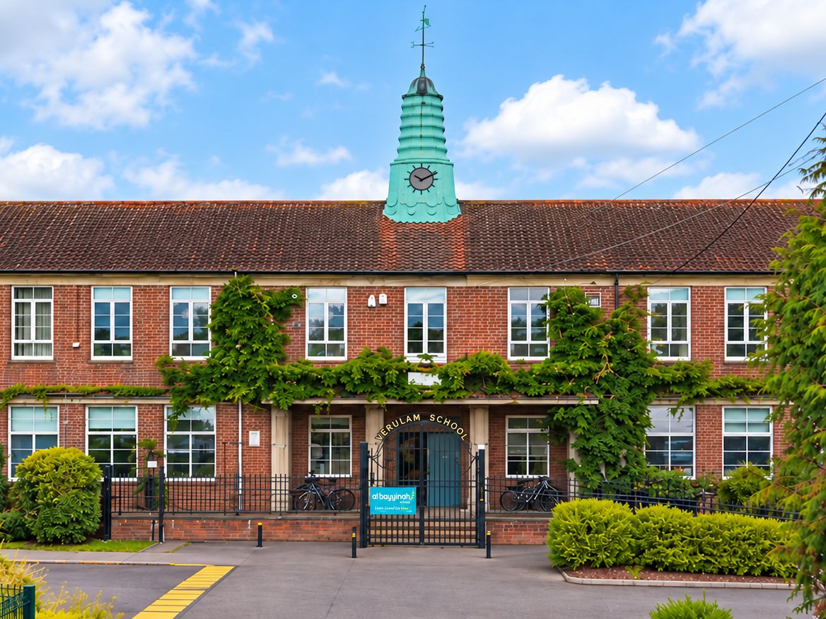 The front of Verulam School: a red-brick building with a green clock tower, with the Al Bayyinah banner on the gate at the main entrance.