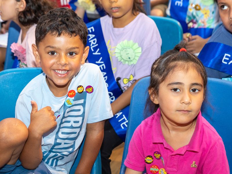 A smiling boy giving a thumbs up, his T-shirt covered in reward stickers, beside a classmate at the early years graduation.